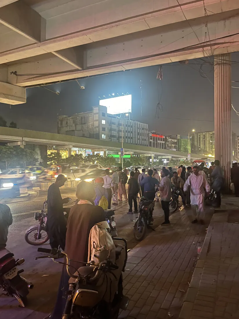 A crowd waits on the footpath beneath a flyover at night, with motorbikes parked across the pavement and traffic passing where riders board.
