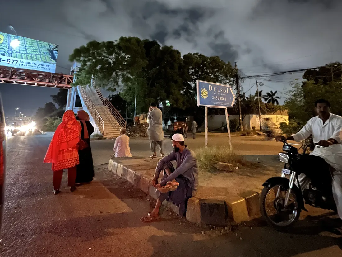 Night at an informal pickup point: people wait on a concrete road median near a pedestrian bridge, with no shelter or seating.