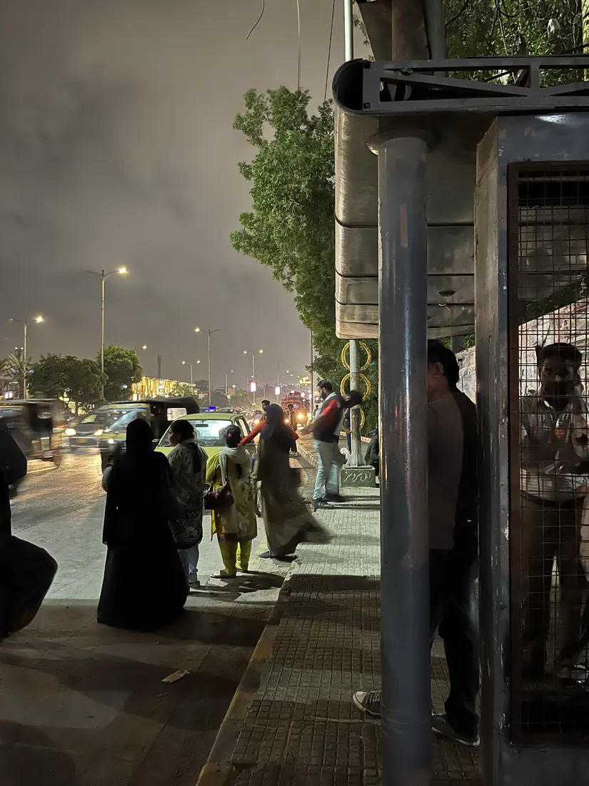 Night in Karachi: a metal roadside shelter occupied by men, while women wait out at the kerb beside moving traffic to flag a rickshaw.