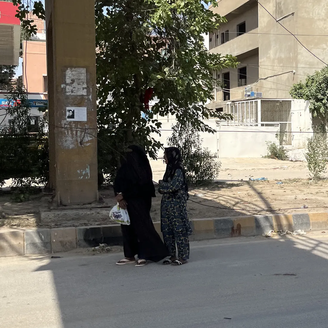 Two women stand waiting beside a flyover pillar in daylight, with no shelter, seating or signage.