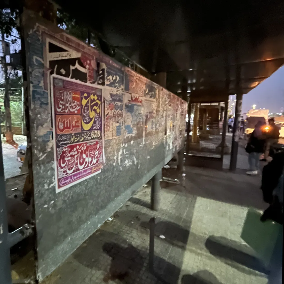 A roadside shelter at dusk with its back panel stripped to bare concrete and covered in layered posters, unlit and empty.