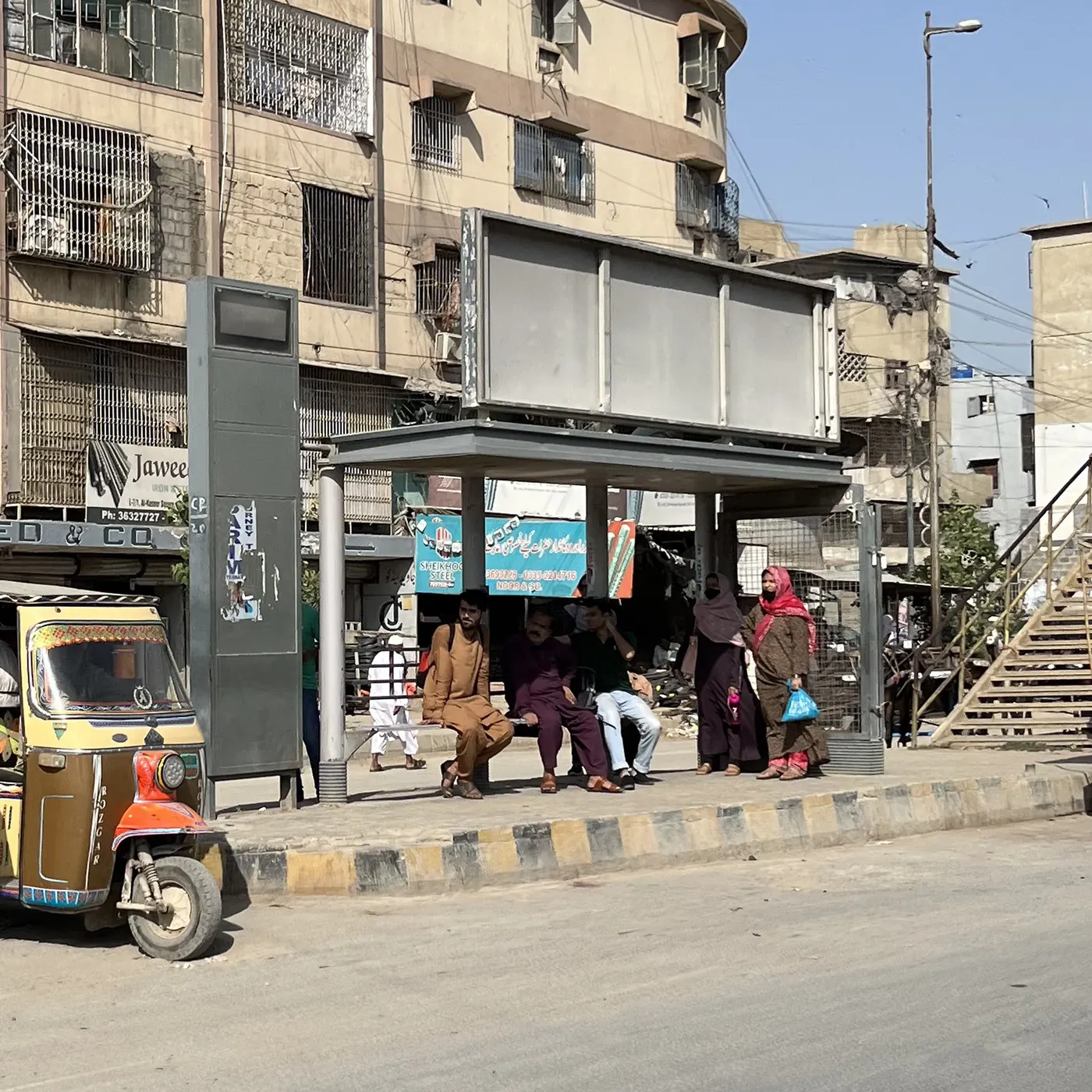 A Karachi bus shelter in daylight: men occupy the full length of the bench while two women stand outside the shelter holding bags, waiting at the kerb.