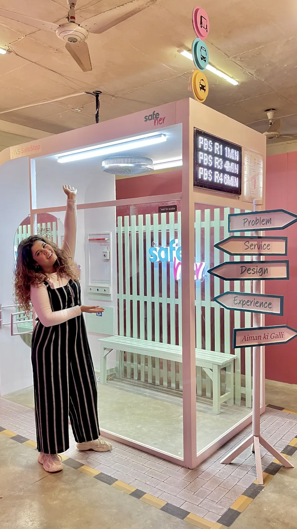 The full-scale SafeStop built for the IVS Degree Show, with its lit canopy, arrivals board, bench and a signpost reading Problem, Service, Design, Experience, seen alongside a visitor for scale.