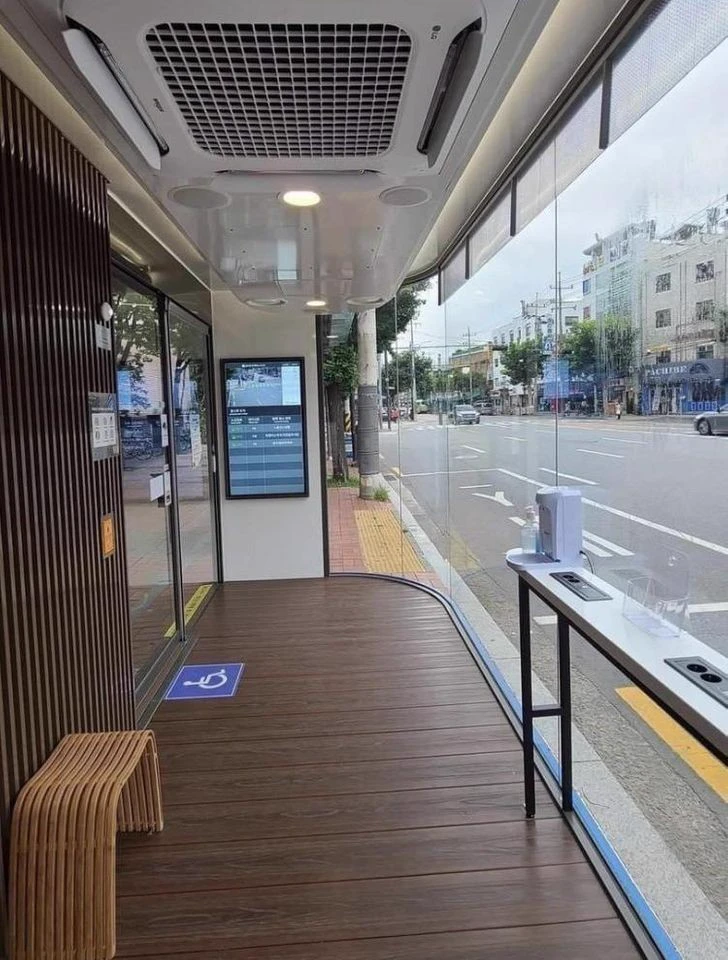 Reference image: a modern glass-walled bus shelter interior with wooden seating and a digital information screen, opening onto a street.